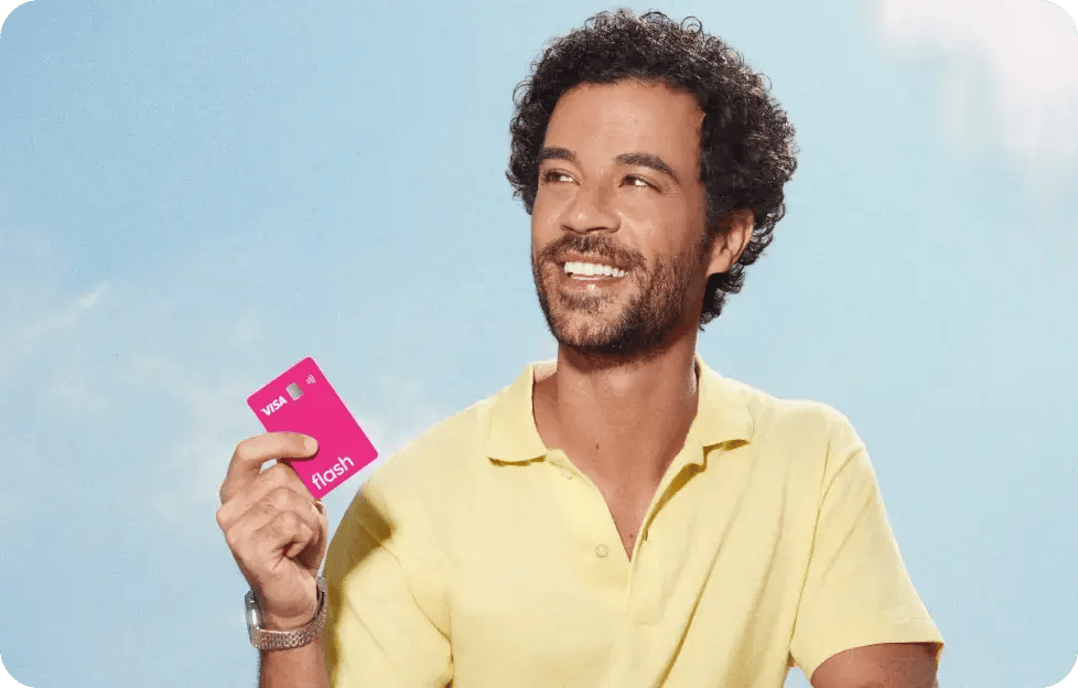 Homem sorrindo segura o cartão multibenefícios da Flash em um ambiente externo com céu azul.